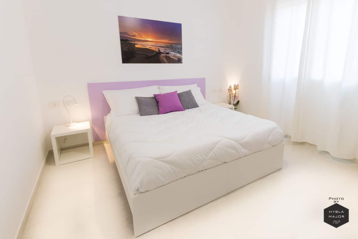 Un dormitorio moderno con una cama blanca, un cabecero morado y un cuadro de atardecer sobre la cama.