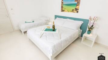 Una cama doble blanca con cabecera color turquesa, una mesita de noche con una botella y un vaso, y una pequeña mesa con una planta.
