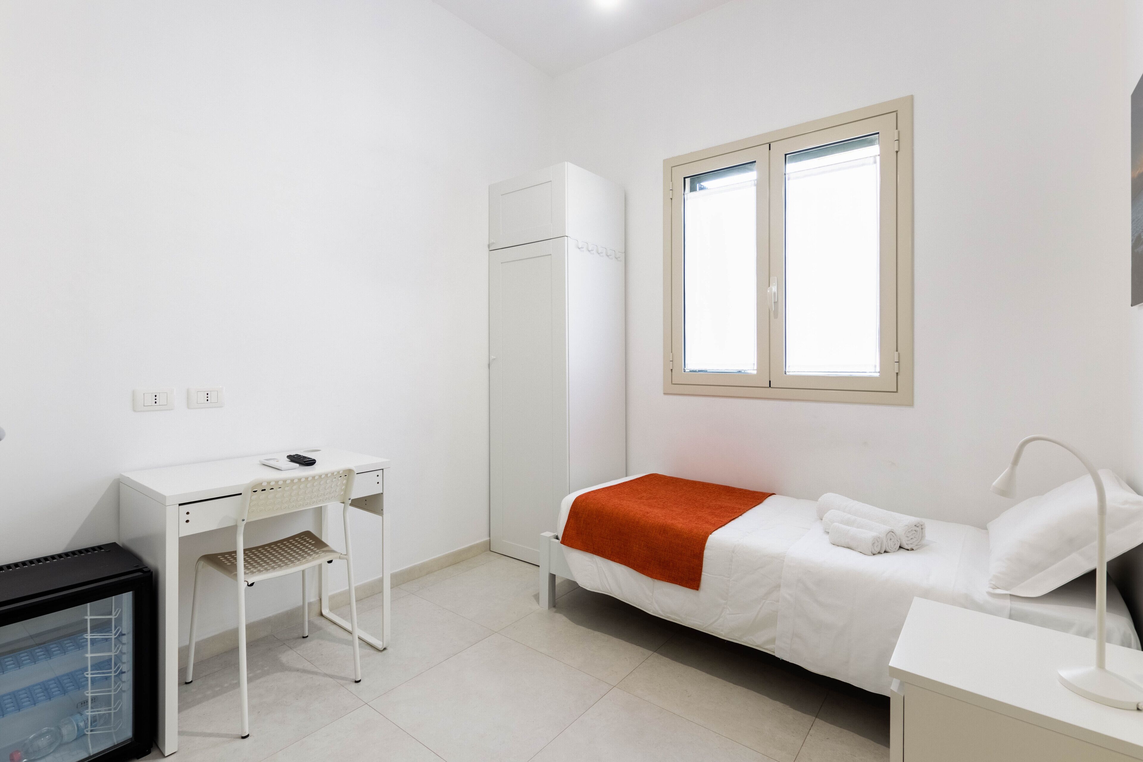A small, clean room with a bed, a desk, a chair, and a refrigerator.