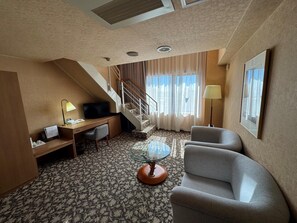 A hotel room with a desk, two armchairs, a glass coffee table, a TV, a lamp, a staircase, and a window with curtains. - Ohta Grand Hotel (Ota)