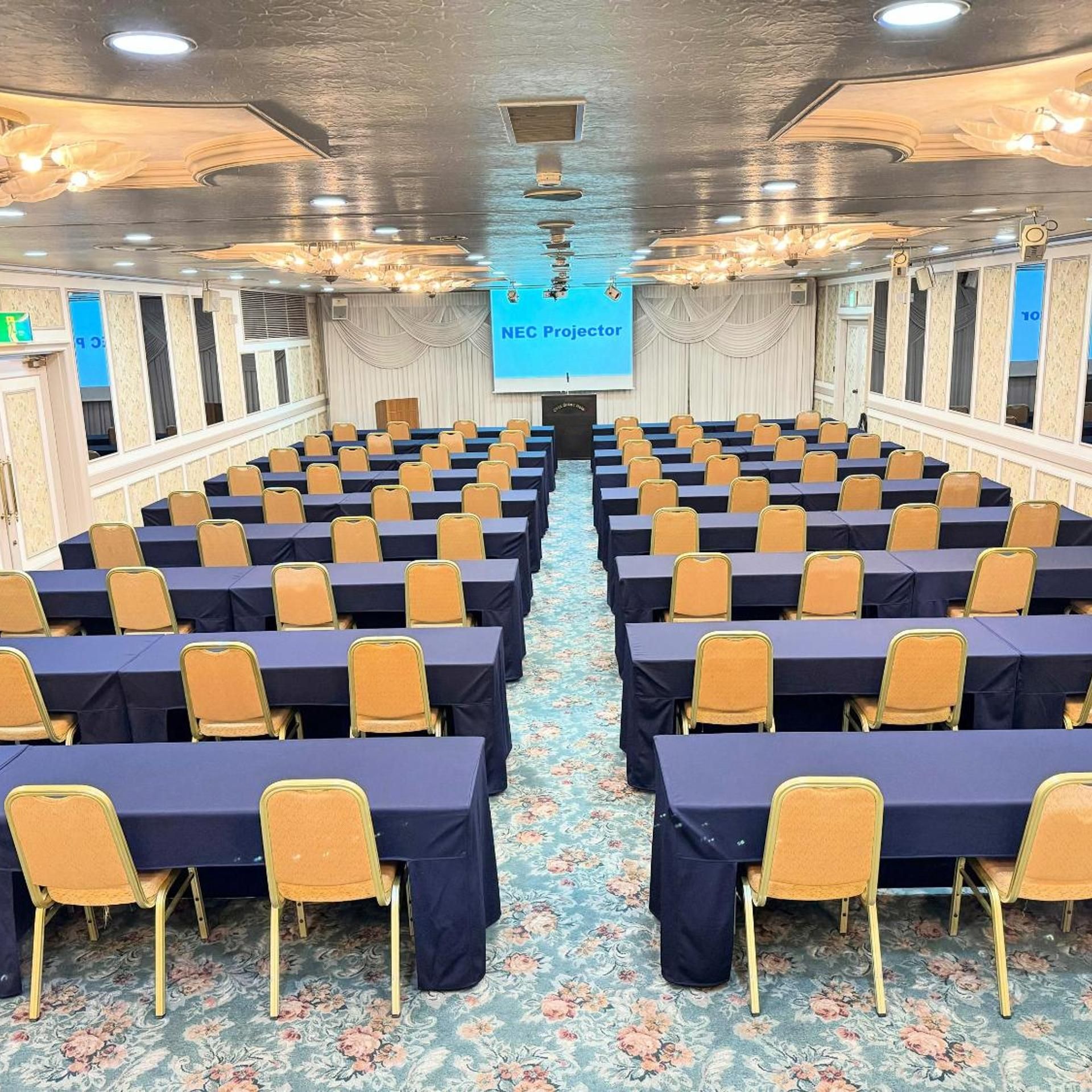 A conference room with rows of tables and chairs, a projector screen, and a floral carpet.