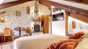 Een slaapkamer met een stenen muur, houten balken, een openhaard, een bed met gestreepte kussens en een televisie.