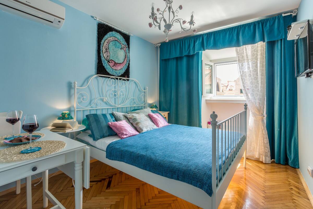 Une chambre à coucher avec un lit, une table de chevet, une table avec des verres de vin et une fenêtre avec des rideaux.