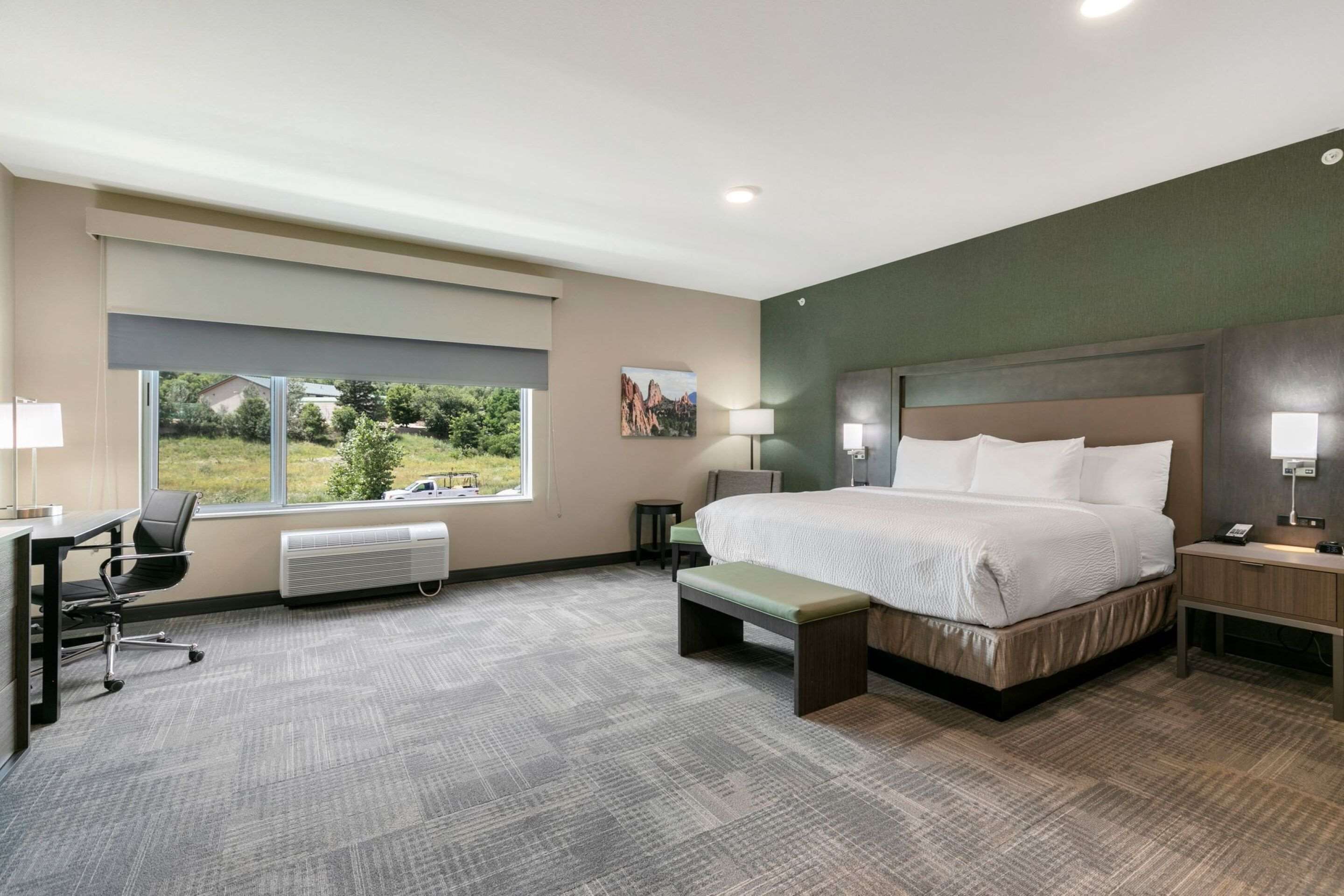 A hotel room with a large bed, a desk, a chair, a bench, a nightstand, and a window with a view of greenery.