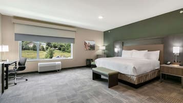 A hotel room with a large bed, a desk, a chair, a bench, a nightstand, and a window with a view of greenery.