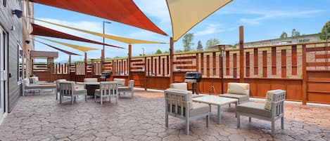 Un patio al aire libre con suelo de piedra hexagonal, mobiliario exterior con sillas y mesas blancas, y grandes estructuras de sombra.
