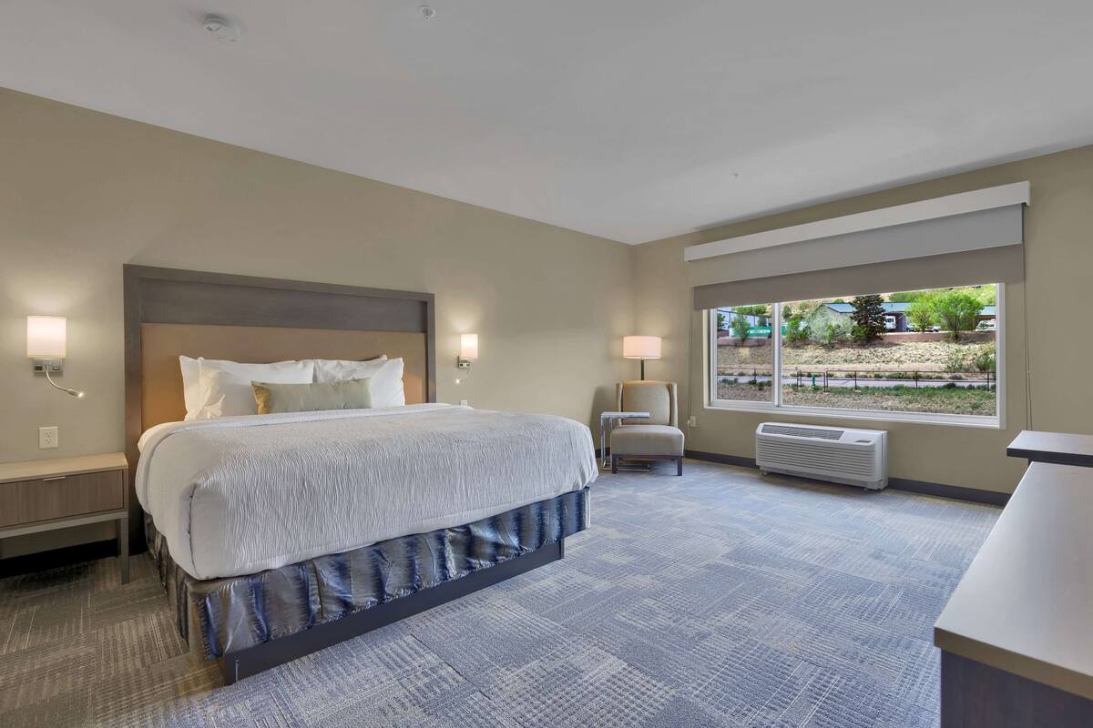 Ein Hotelzimmer mit einem großen Bett, Nachttischlampen, einem Sessel, einem Schreibtisch und einem Fenster mit Blick ins Freie.