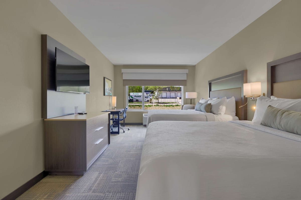 Ein Hotelzimmer mit zwei Betten, einem Schreibtisch, einem Sessel, einem Fernseher und einem Fenster mit Ausblick.