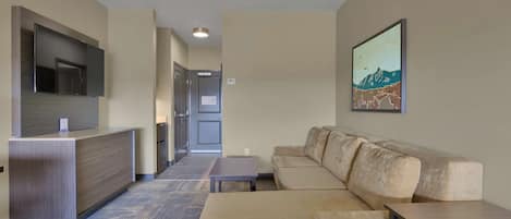 Quarto de hotel moderno com sofá, uma mesa pequena, televisão de tela plana presa a um armário de madeira e um quadro na parede.