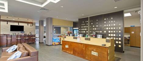A modern hotel lobby with a reception desk, a seating area with a sofa, a kitchen area, and a large TV screen.