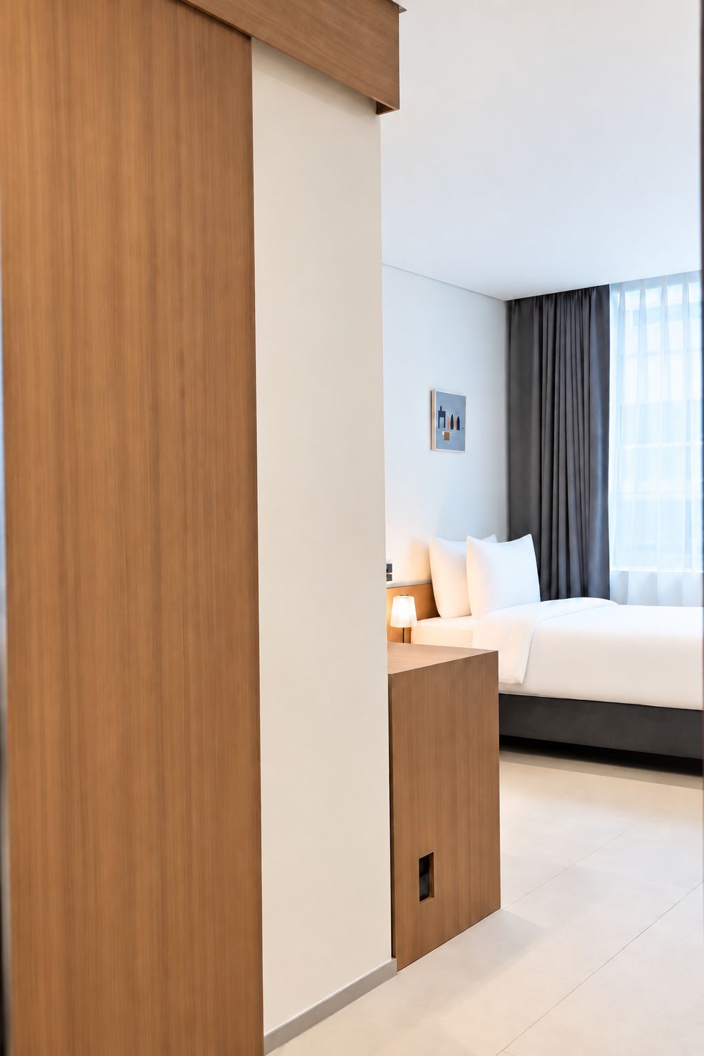 A modern hotel room with a large bed, a bedside table, a lamp, and a framed picture on the wall.
