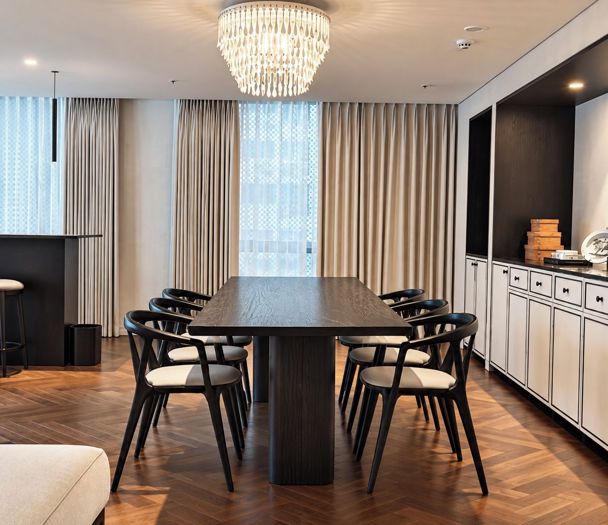 A modern dining area with a large rectangular table, six chairs, a chandelier, and a kitchen area with cabinets and a countertop.