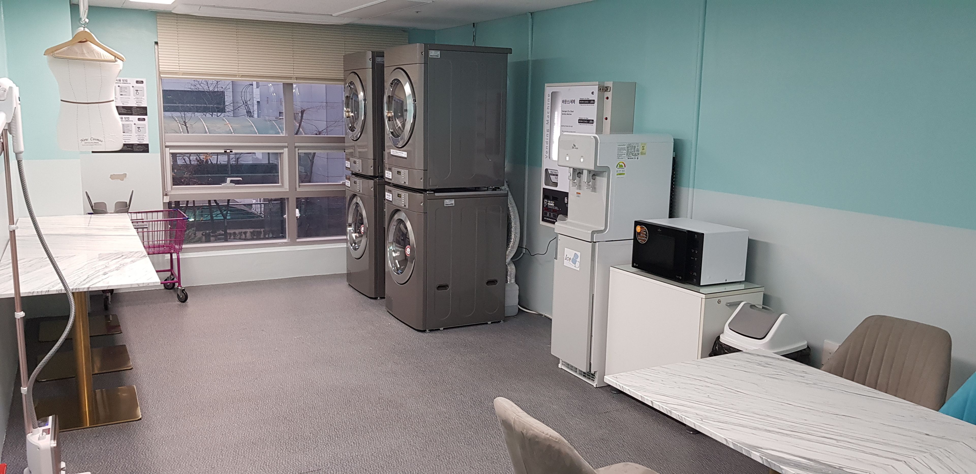A clinical room with a bed, a desk, a chair, a water dispenser, a microwave, and a window with blinds.