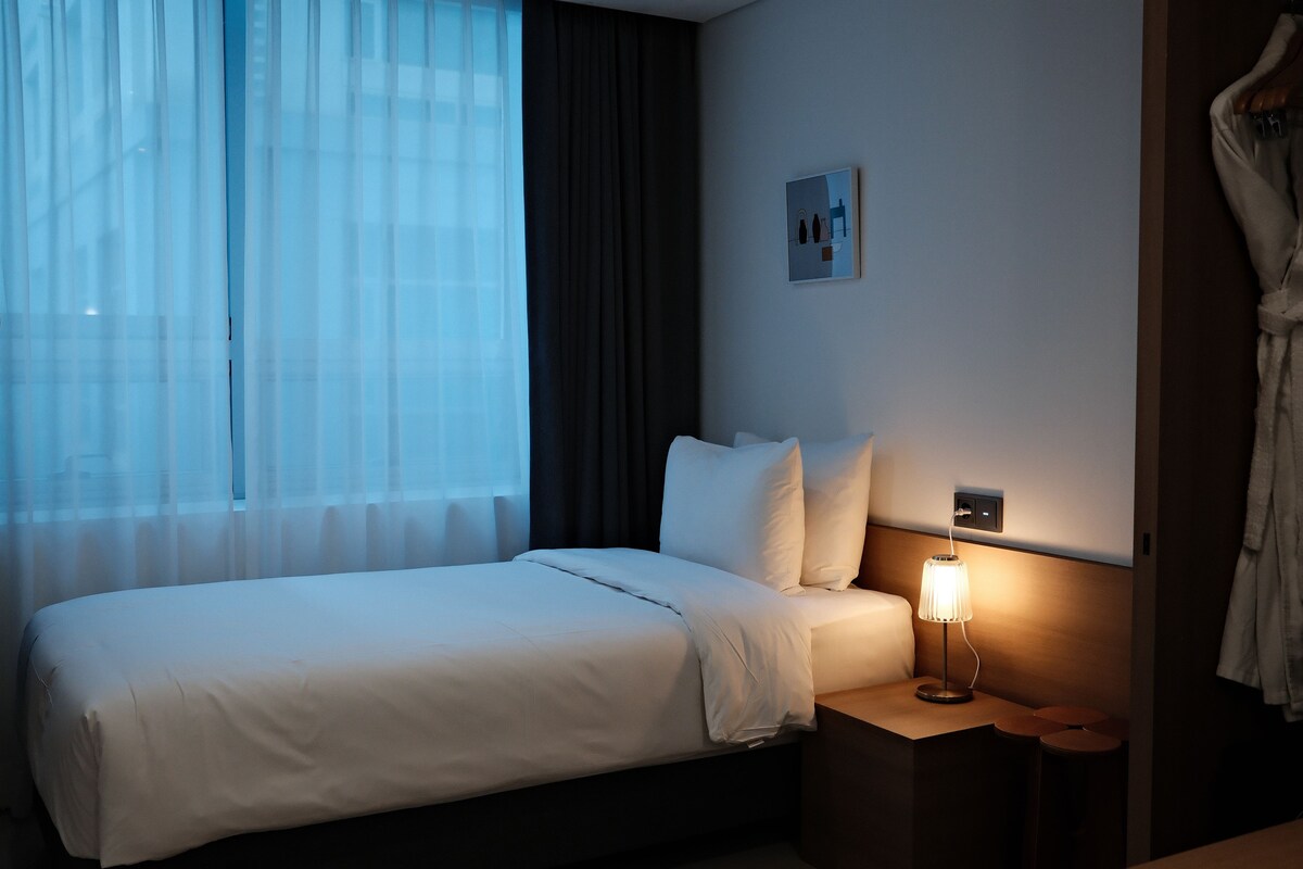 Une chambre d’hôtel avec un lit, une table de chevet avec une lampe, une prise de courant murale et une fenêtre avec des rideaux légers.