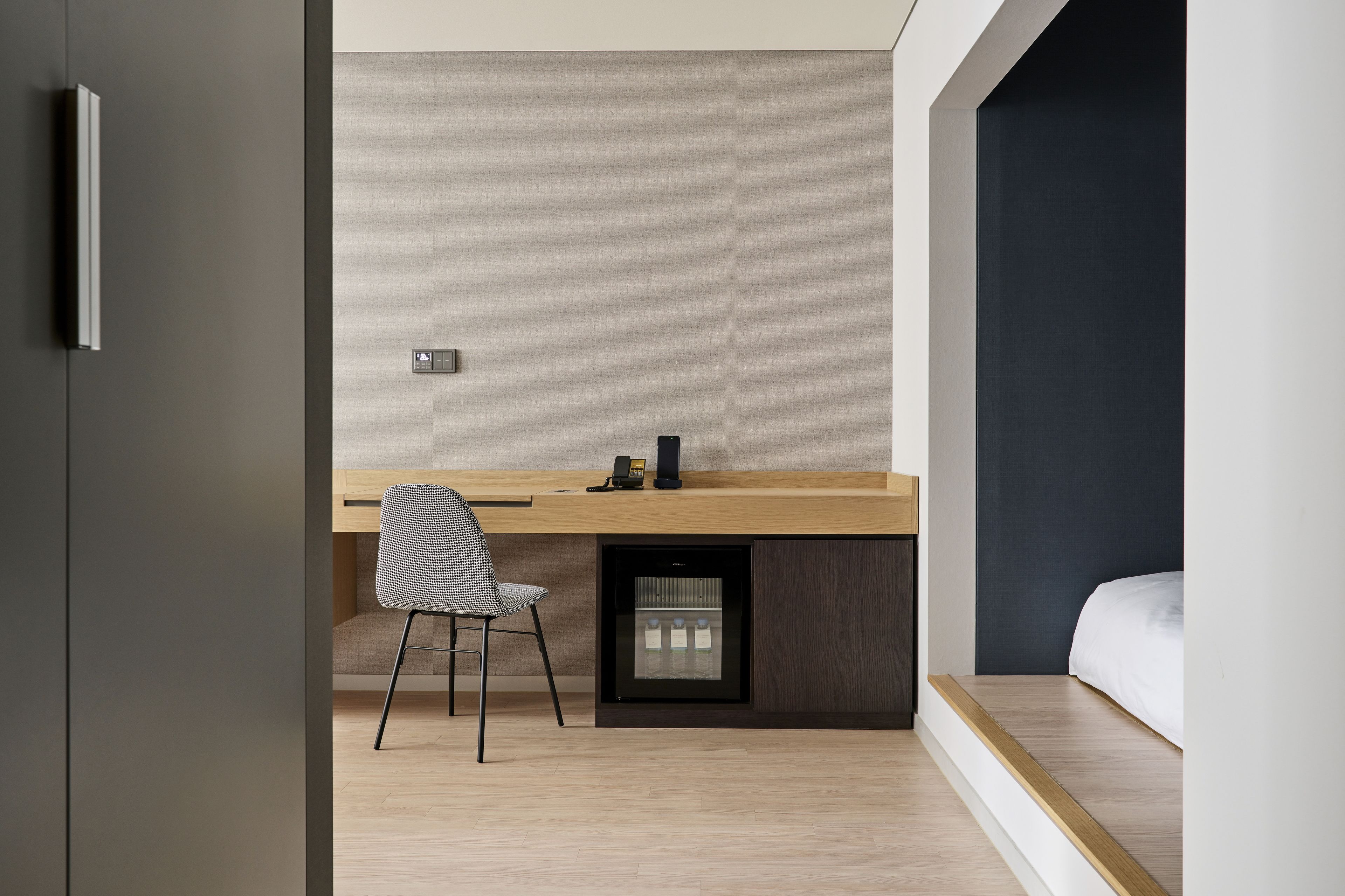 Una habitación de hotel moderna con un escritorio de madera, una silla, una cama y un refrigerador empotrado pequeño.