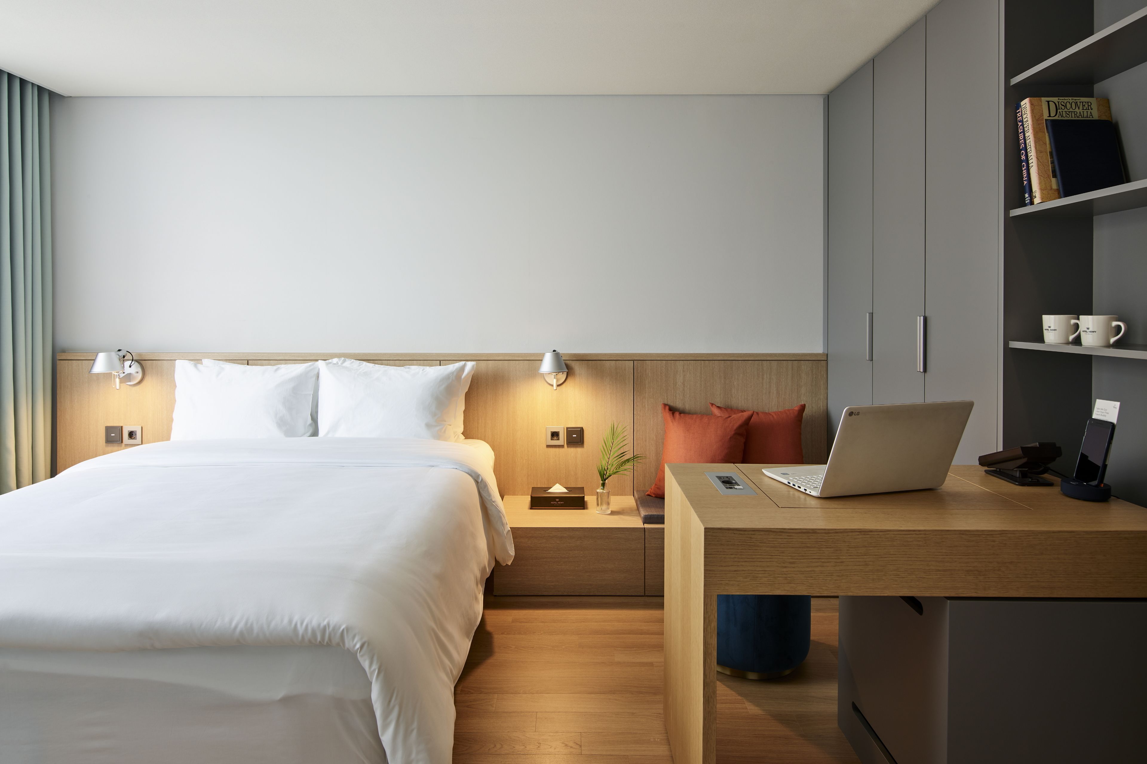 Una habitación de hotel moderna con una cama bien hecha, un escritorio de madera con una computadora portátil, una estantería y una mesita de noche con una lámpara.