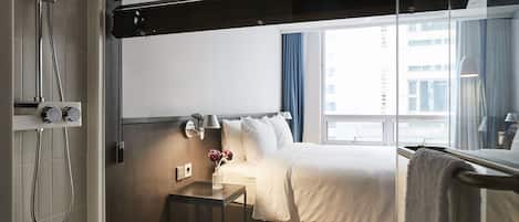 Habitación de hotel con cama, mesita de noche, ducha y ventana con vista a edificios.