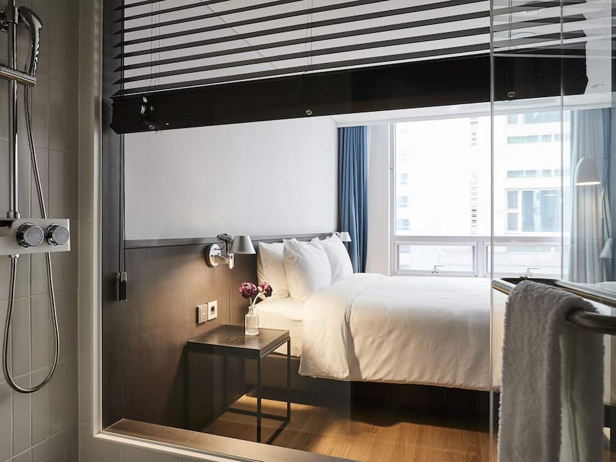 Habitación de hotel con cama, mesita de noche, ducha y ventana con vista a edificios.