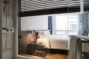 A hotel room with a bed, a bedside table, a nightstand, a shower, and a window with a view of buildings.