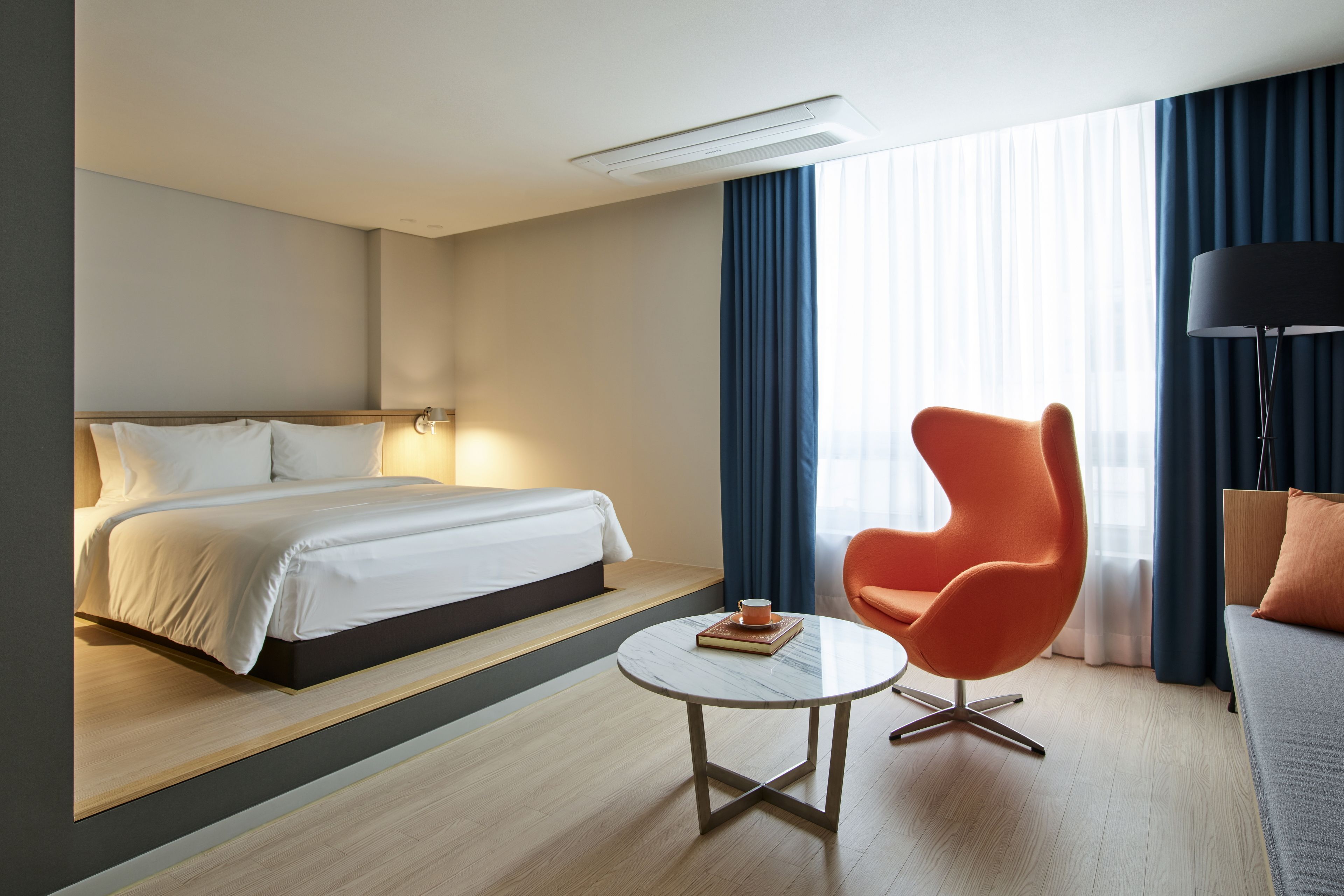 A hotel room with a bed, a round table, a modern orange chair, a sofa, and a lamp.