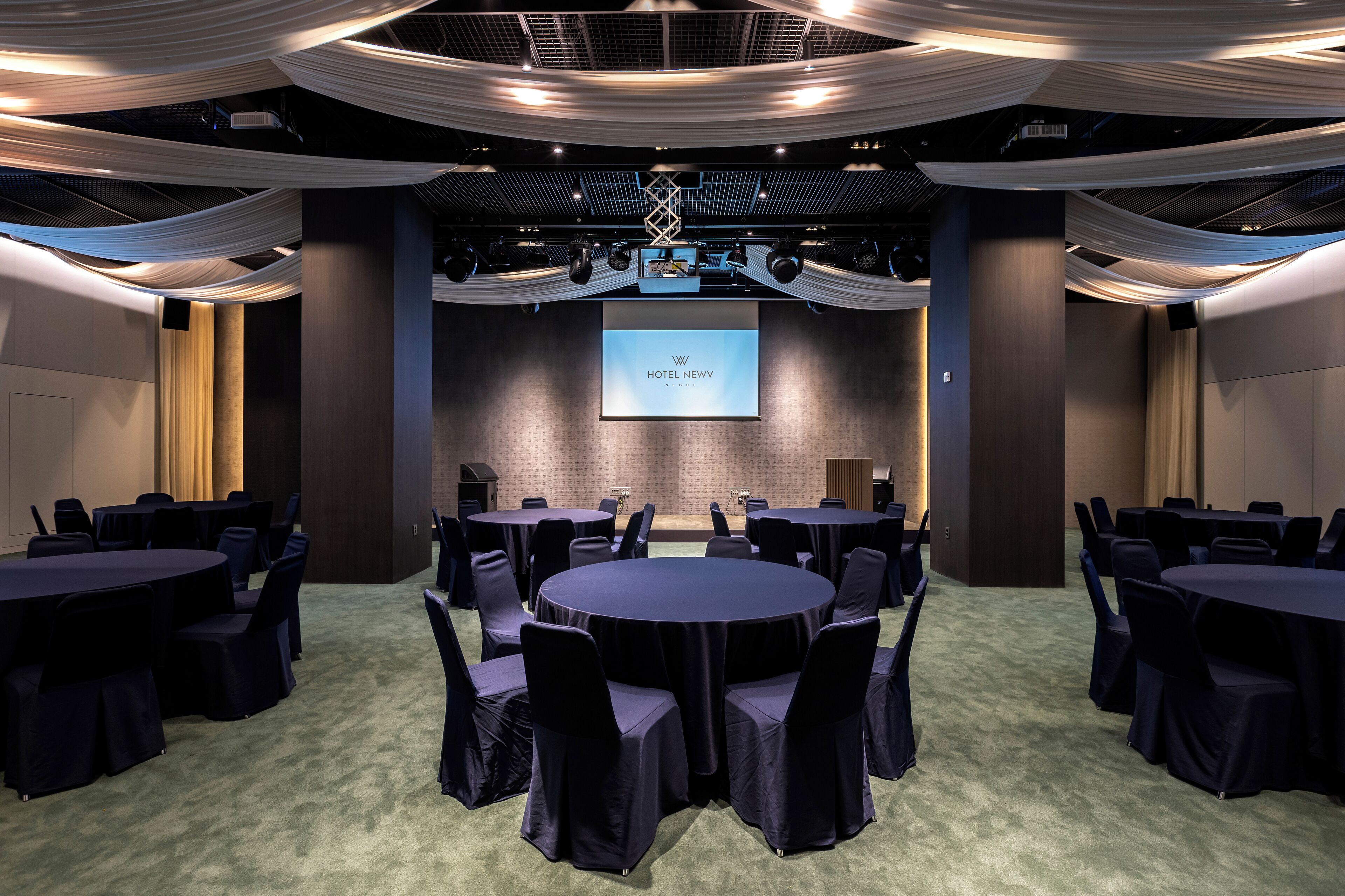 Un salón de conferencias con mesas y sillas redondas, una pantalla de proyector y telas decorativas en el techo.