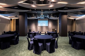Une salle de conférence équipée de tables et de chaises rondes, d’un écran de projection et de panneaux de tissu décoratifs suspendus au plafond.
