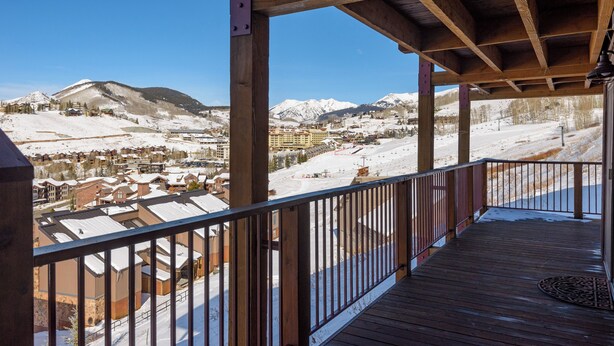 A wooden deck with a metal railing overlooking a snowy mountain town with buildings and a clear blue sky. - Ski-In/Out! 3/2 Slopeside Spacious Condo w/ Amazing Mtn Views!! (Mt. Crested Butte)