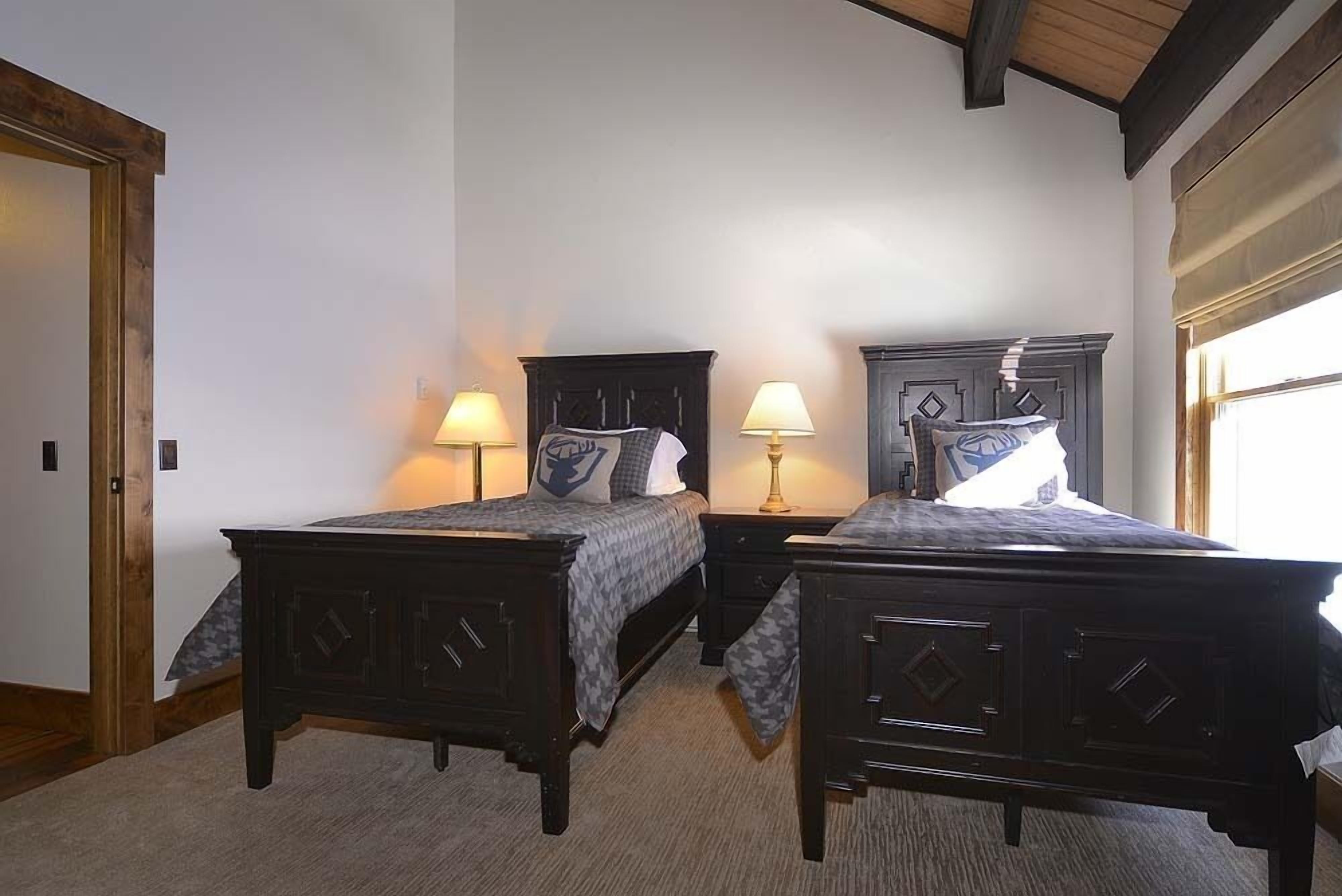 A bedroom with two dark wooden beds, each with a matching nightstand and lamp. The room has a wooden ceiling and a window with a view.