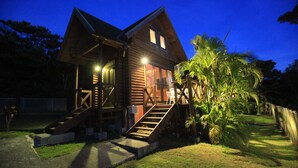 A wooden house with a staircase leading to the front door, surrounded by palm trees and a well-maintained lawn. - Tokashiki Guest House (Tokashiki)