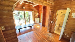 A wooden cabin interior with a bench, wooden floors, a bathroom with a toilet, and a window with a view of greenery. - Tokashiki Guest House (Tokashiki)