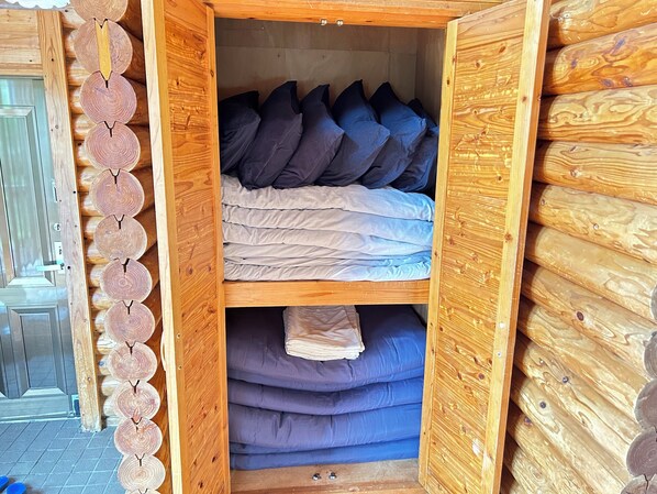 A wooden cabin interior with a bed, pillows, and a shelf with folded blankets. - Tokashiki Guest House (Tokashiki)