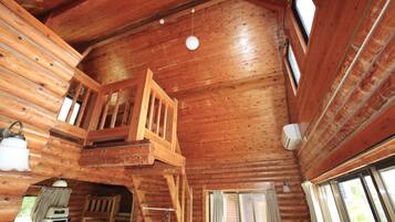 A cabin interior with wooden walls, a loft bedroom with a wooden ladder, a kitchen area, and a large window providing natural light.