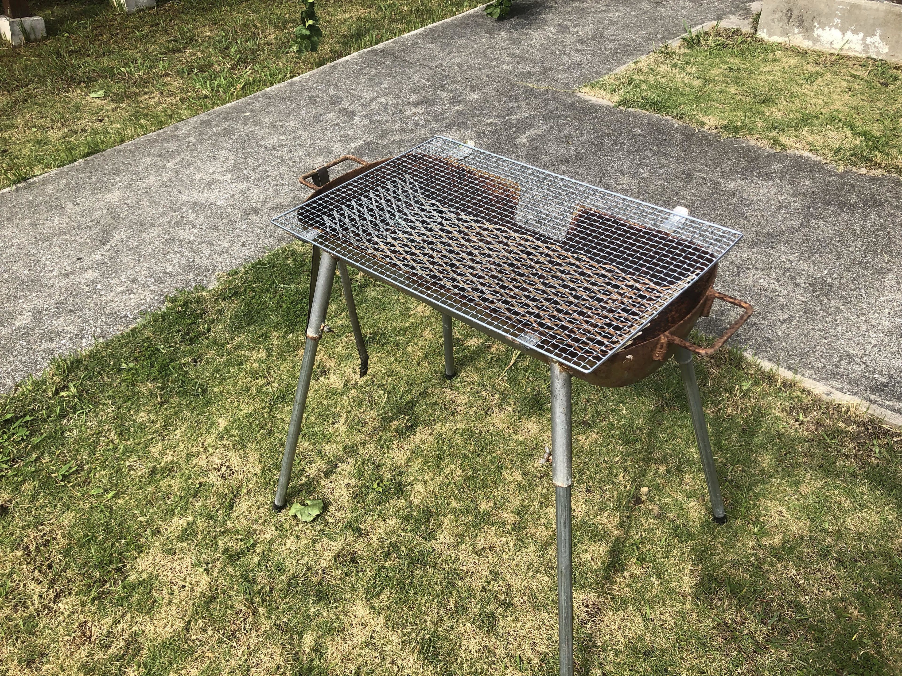 A metal grill table with a mesh top, set on a grassy area with a concrete path nearby.