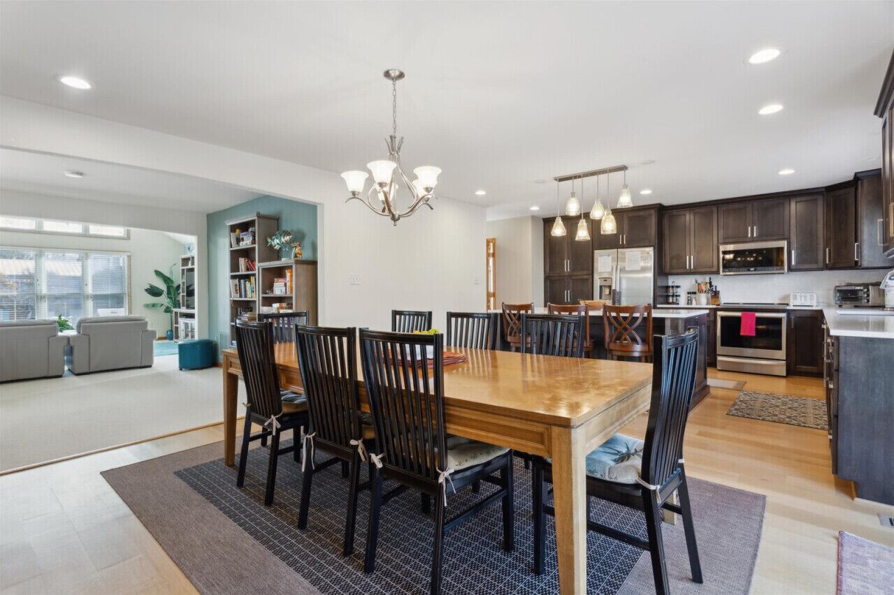 A spacious living room with a large wooden dining table, dark wooden chairs, a chandelier, and a bookshelf.