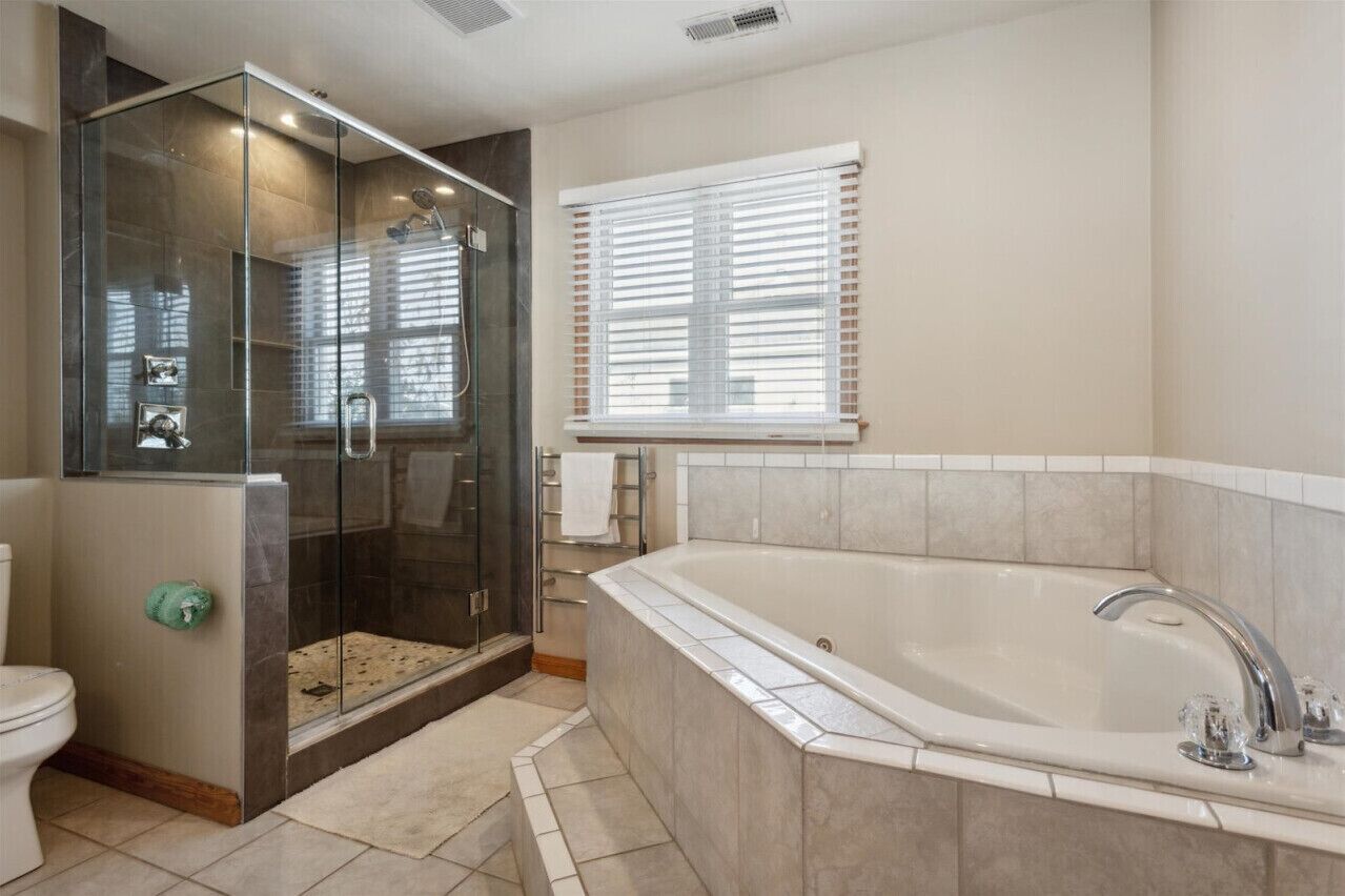 A bathroom with a glass-enclosed shower, a large bathtub, and a window with blinds.