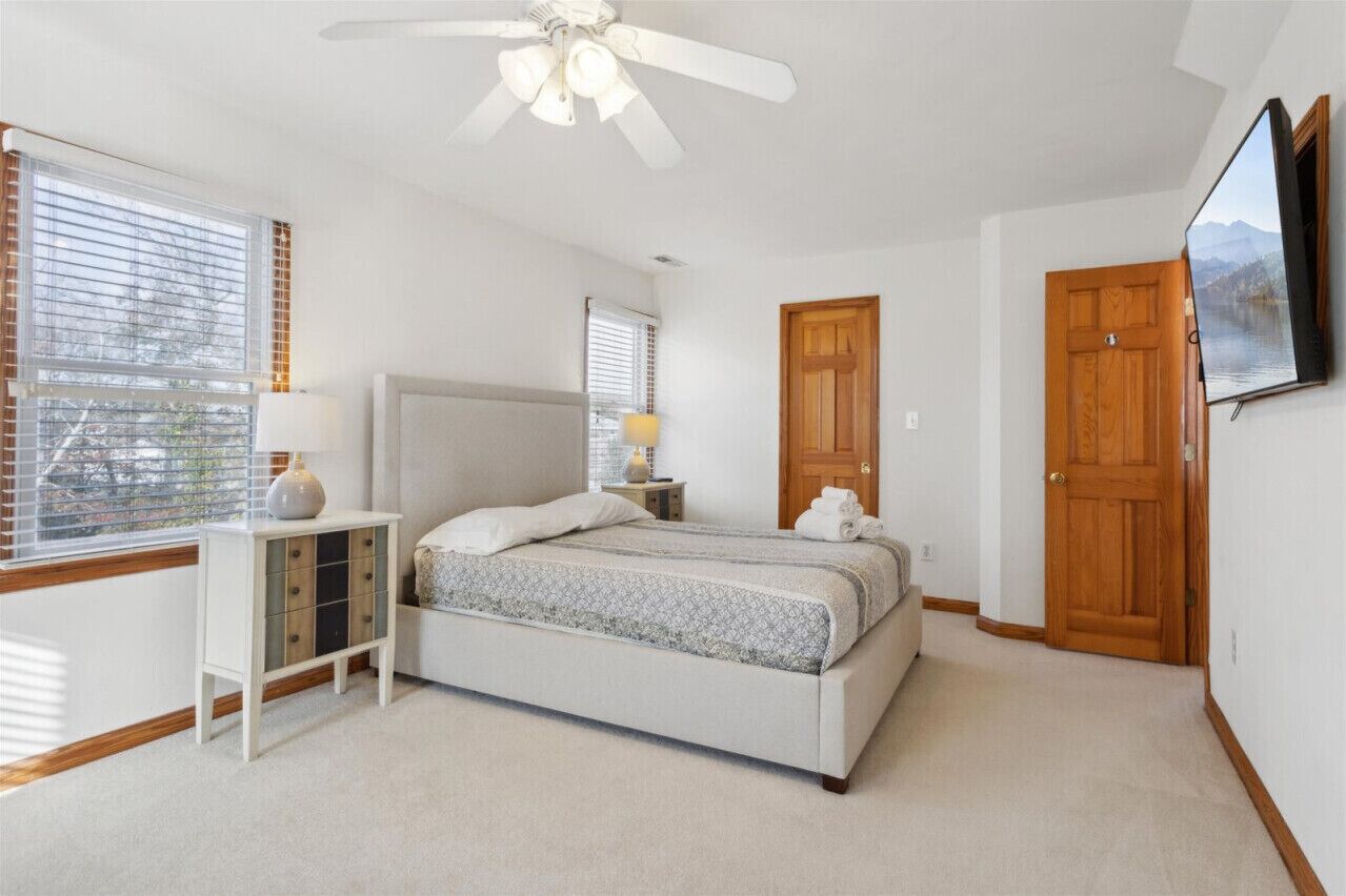 A bedroom with a bed, a nightstand, a ceiling fan, a TV mounted on the wall, and two doors.
