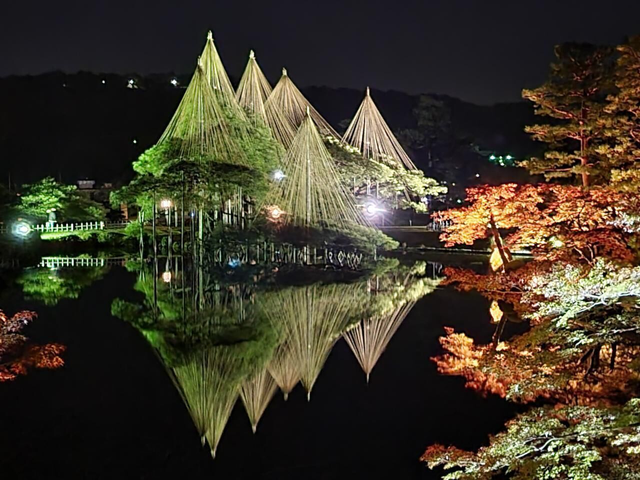 A traditional Japanese structure with a reflection in the water, surrounded by trees and a fence.