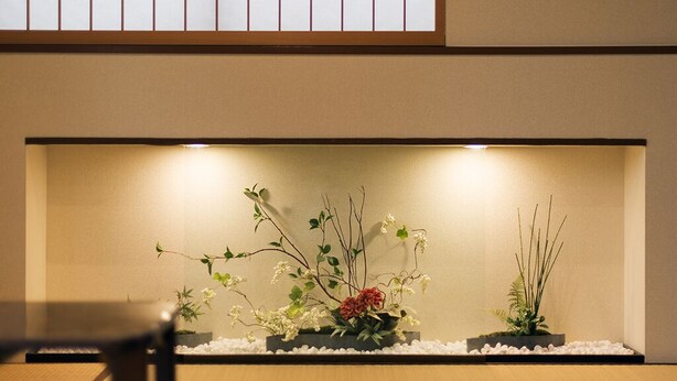 Ein minimalistischer Innenraumgarten mit drei Pflanzgefäßen, in denen unterschiedliche Pflanzen und Blumen stehen, vor einer neutralen Wand mit einem Holzbalken darüber.