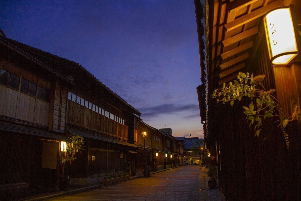 A narrow street with traditional wooden buildings, illuminated by warm lighting, and a clear sky.