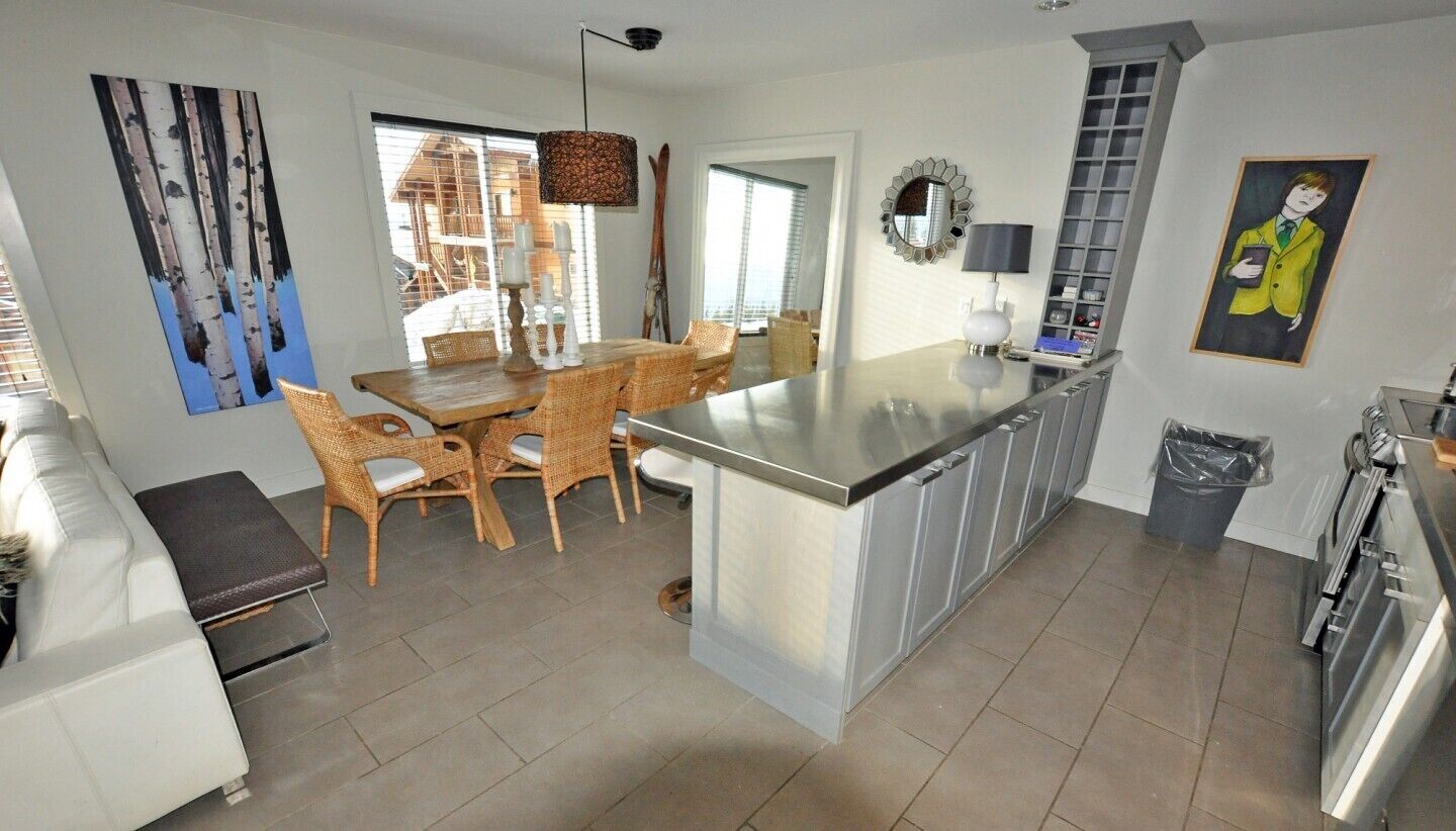 A modern kitchen with a central island, dining area with wicker chairs, and a large window providing natural light.