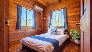 Ein Schlafzimmer einer Holzhütte mit einem Bett, zwei mit Vorhängen versehenen Fenstern, einer Klimaanlage und einer Topfpflanze.