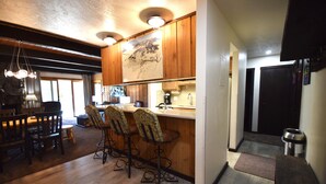 A kitchen with wooden cabinets, a bar area with stools, a stone fireplace, and a dining area with wooden tables and chairs. - Great Location=Great Vacation. Dog Friendly Condo w/ Awesome Ski Access (Mt. Crested Butte)