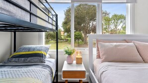 A bedroom with bunk beds, a bedside table with a lamp, and a window with a view of trees and buildings. - RYE Classic- 4bed, perfect location-up to 13 guests, foxtel- BONUS* NIGHTS OFFER (Rye)