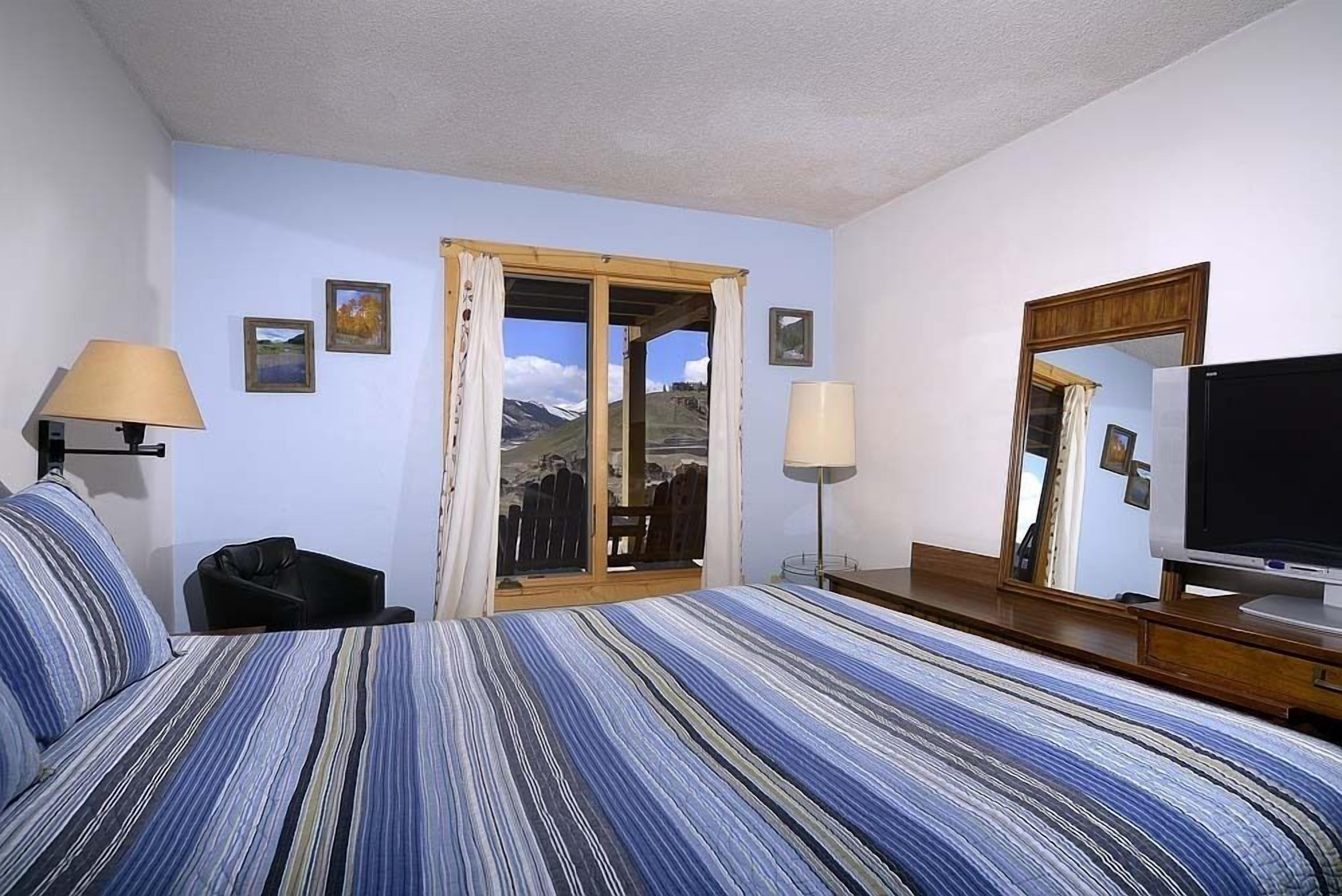 A hotel room with a bed, two lamps, a television, a mirror, and a door leading to a balcony with a mountain view.