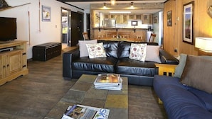 A living room with a sofa, coffee table, and TV. There is a kitchen in the background. - Close to Everything! Ski-In/Out Condo. Doggie Friendly, Mtn Views, Fireplace (Mt. Crested Butte)