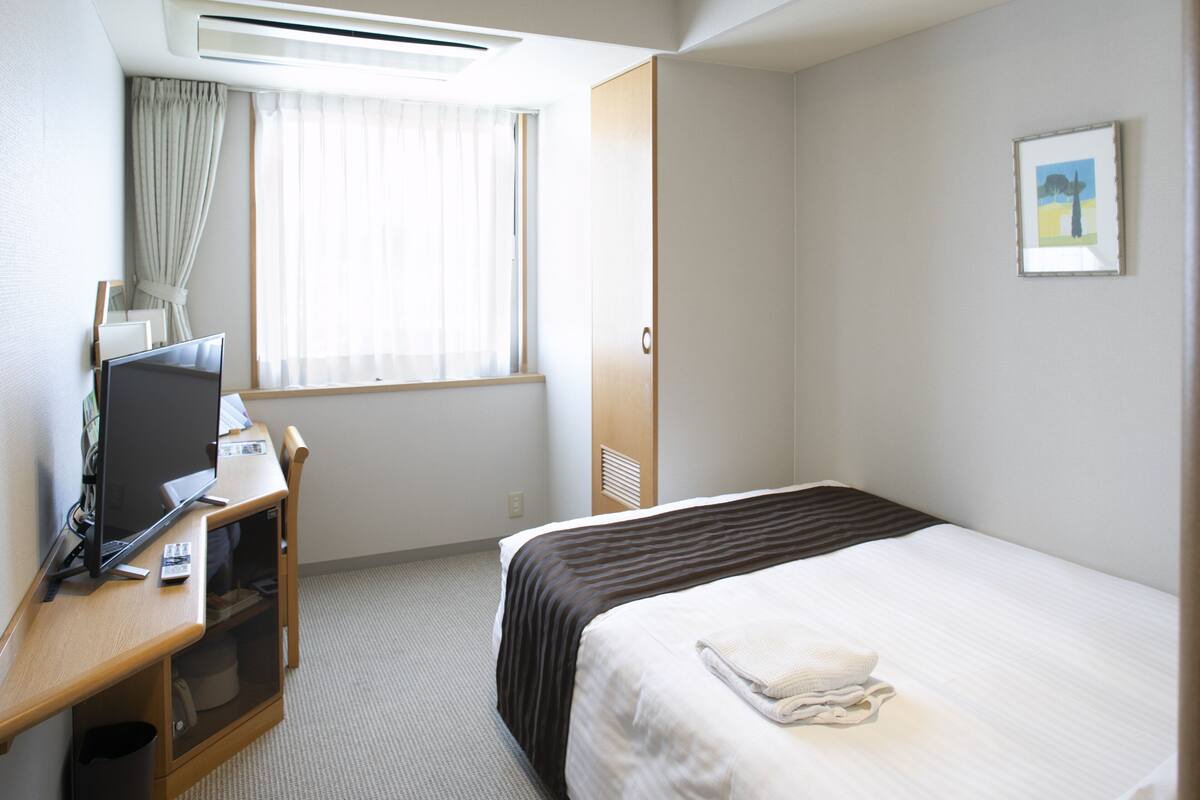 A hotel room with a bed, a desk with a computer, a window with curtains, and a framed picture on the wall.