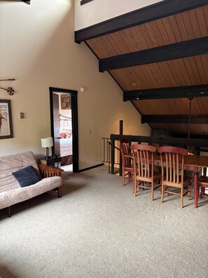 A cozy room with a high ceiling, a wooden dining table with chairs, a sofa, and a lamp.