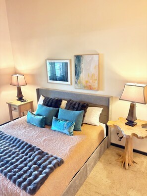 A hotel room with a bed, two bedside tables, a nightstand, a lamp, and two framed artworks on the wall.