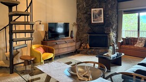 A living room with a stone wall, a TV, a fireplace, a yellow armchair, and a staircase. - Spacious Mountain Condo Retreat! Ski-In/Out w/ Views, Fireplace, Dog Friendly (Mt. Crested Butte)