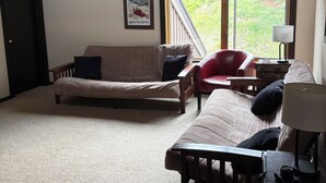 A spacious living area with a high wooden ceiling, a large window with a view of greenery, a red chair, a beige sofa, and a small table with a lamp. - Spacious Mountain Condo Retreat! Ski-In/Out w/ Views, Fireplace, Dog Friendly (Mt. Crested Butte)